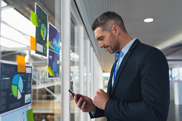 Mature adult man in suit standing next to glass wall tapping smartphone with charts, sticky notes