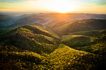 Aerial view of breathtaking sunrise over rolling mountains covered in mix of lush green and autumnal foliage, with dramatic clouds streaking across sky, casting warm, golden light over the landscape.