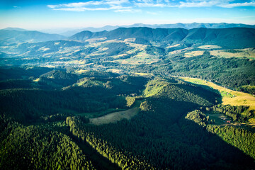 Aerial view of densely forested mountains stretching into distance under clear blue sky with scattered clouds. Landscape reveals valleys and distant fields, showcasing natural beauty of region.