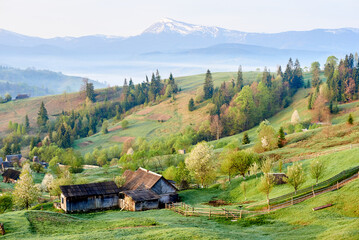 Serene morning landscape with rural village nestled among rolling hills. Wooden barns and blossoming trees dot foreground, while mist blankets valleys. Snow-capped mountains rise in background.