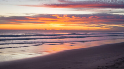 Dramatic sunset colors the sky and reflects on wet sand, creating a stunning coastal scene.