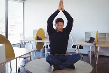 Pacific Islander man wearing navy sweater meditating on round table in open-plan office with laptop