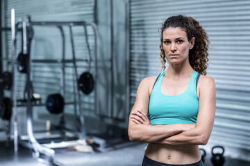 Female trainer standing arms crossed in gym by squat rack wearing workout attire, copy space