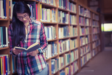Student leaning against tall wooden bookshelf in library aisle, reading open book with glasses