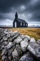 Black wooden church under dramatic stormy sky with snow-covered mountains in the distance and a stone wall in the foreground on the Icelandic countryside