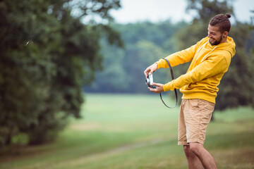 Fototapeta premium Man wearing yellow hoodie and tan shorts holding film camera smiling in grassy meadow, copy space