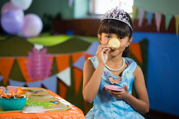 Girl sipping from yellow toy teacup with pink saucer at party table near pastel balloons