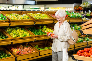 Senior woman examining red tomatoes in produce section holding plastic shopping bag, copy space