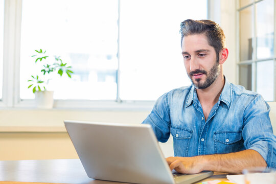Fototapeta Man typing on silver laptop at wooden desk in bright office with potted plant, copy space