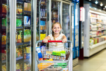 Girl pushing shopping cart loaded with eggs, cereal box and beverage cartons along grocery aisle