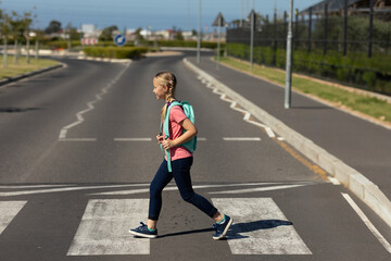 School-age girl crossing two-lane road on zebra crosswalk near shrubs, carrying mint green backpack © wavebreak3