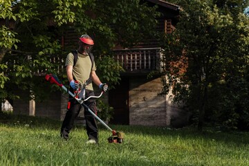 Man using a trimmer to maintain grass near a wooden house during a sunny day