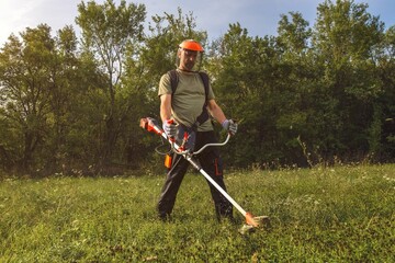 Person using a string trimmer in a grassy area surrounded by trees during daylight