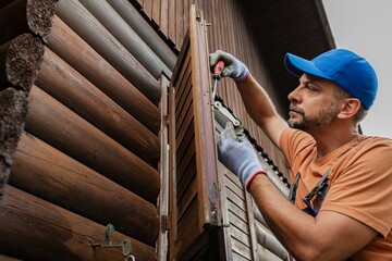 Worker repairs wooden shutters on rustic cabin during overcast afternoon