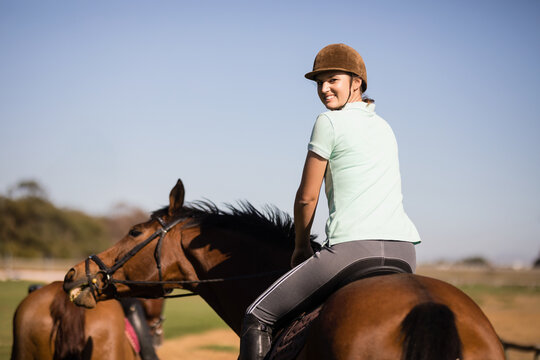 Fototapeta Female equestrian riding brown horse in sunny paddock wearing helmet and holding reins
