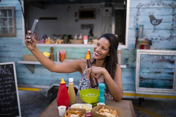 Woman taking selfie at wooden table in front of food truck holding beer bottle and smartphone