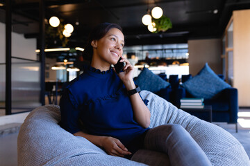 Asian woman sitting in office lounge holding smartphone to ear beside blue sofa with pendant lights
