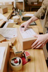Person packing parcels on rustic wooden table with various shipping supplies and open laptop in the...