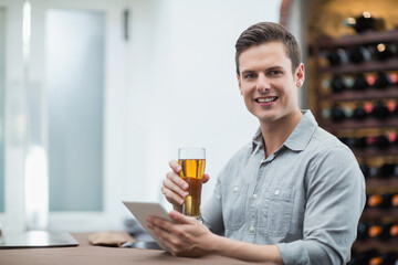 Man wearing grey shirt sitting at bar counter holding beer glass, using tablet by wine rack