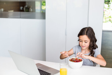 Girl sitting in home kitchen eating cereal holding spoon with laptop and orange juice, copy space © WavebreakMediaMicro