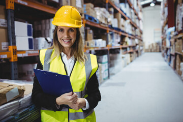 Female warehouse supervisor standing in aisle wearing yellow safety helmet and vest, copy space