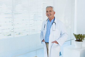 Senior doctor standing in clinic with white lab coat, stethoscope, blinds and plant, copy space