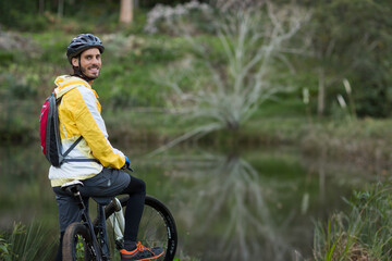 Man smiling astride mountain bike at forest pond edge wearing rain jacket and helmet, copy space