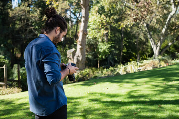 Fototapeta premium Male photographer adjusting DSLR camera on grass lawn in park with sunlight filtering through trees