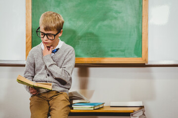 School-aged boy wearing eyeglasses is reading book beside stack of books, in front of chalkboard