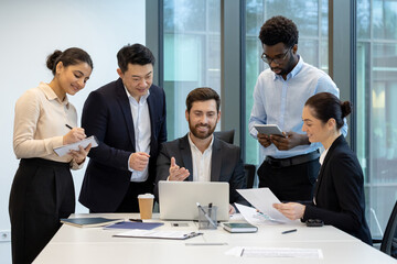 A diverse group of business professionals collaborates around a table in a modern office setting, focused on a laptop and documents.