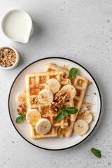 Belgian waffles with banana, nuts and honey on plate served with mint leaves on white table background. Sweet breakfast concept