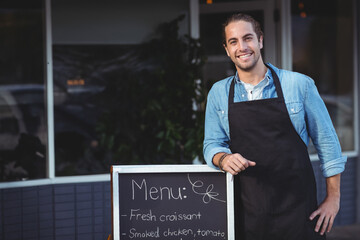 Staff wearing denim shirt and apron leaning on chalkboard outside cafe windows reflecting foliage