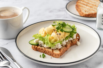 Scrambled eggs on grilled toast with salad leaves, cheese and micro greens on plate on white marble background. Delicious breakfast or brunch
