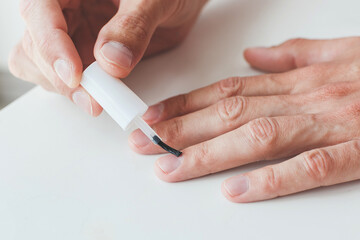 close-up of a man s hands applying clear nail polish or base coat to his fingernails. The hands rest on a white surface, and the focus is on the careful grooming or self-care routine.