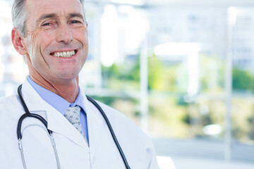 Senior male doctor standing beside window in clinic wearing white coat with stethoscope, copy space
