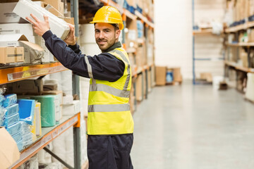 Male warehouse worker wearing hard hat, vest reaching for box on metal rack in warehouse aisle