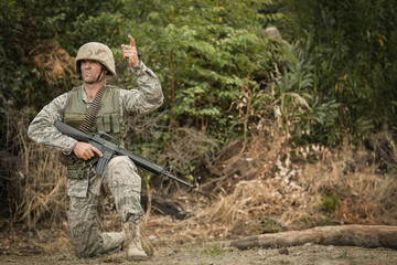Adult male soldier kneeling on one knee in forest edge holding assault rifle, pointing, copy space