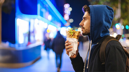 A person in a blue hoodie enjoys a freshly wrapped pita while strolling through a bustling city at night. Vibrant neon lights illuminate the background, highlighting themes of street food culture and