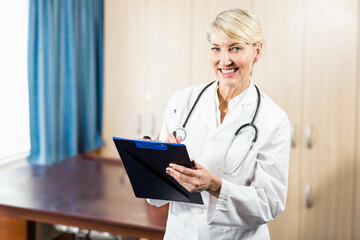 Senior female doctor writing on blue clipboard with pen and wearing stethoscope in clinic office