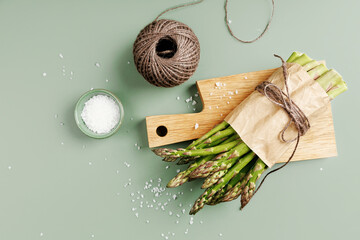 Overhead view of asparagus tips on wooden board with twine and ceramic bowl of salt on green surface