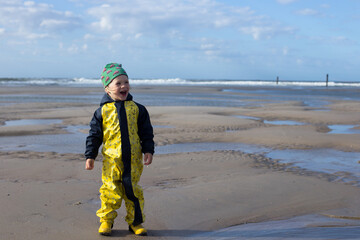 Child in yellow waterproof suit walking on a windy beach near the sea on a cloudy day.