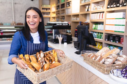 Female bakery attendant holding wicker basket of breads inside bakery shop near espresso machine