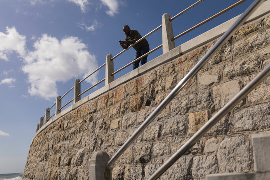 African American man standing on seawall checking smartphone by posts and railings, copy space