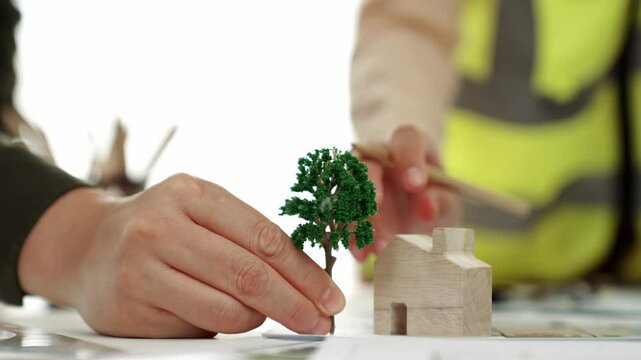 A video of architect using tree and home model to measure blueprint. Architect designing house construction on a table at studio, with architectural plan scattered around. Focus on hand. Delineation.