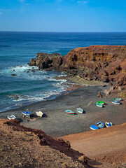 Small, colourful fishing boats rest on the black sand in small bay hidden among red cliffs that rise above blue Atlantic Ocean. Picturesque landscape with unique charm and volcanic beauty in Lanzarote
