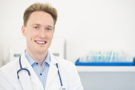 Male doctor standing and smiling in clinical exam room wearing lab coat and stethoscope, copy space