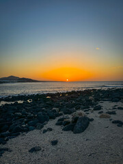 Evening coastal landscape with rippling Atlantic Ocean and distant volcanic mountains. Setting sun bathes sky in warm orange and soft blue hues, illuminating vast stretch of rocky and sandy coastline.