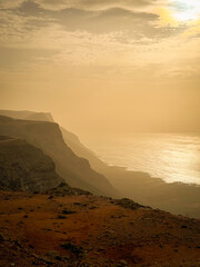 SILHOUETTE: Dramatic coastal cliff disappears into the sunlit, misty ocean. Shimmering water contrasts with dark, layered landforms to create powerful and atmospheric landscape under soft, cloudy sky.