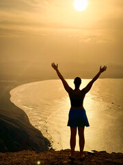 SILHOUETTE, LENS FLARE: Woman with raised arms overlooks a sunlit bay, with shimmering water reflecting soft glow of sky. Dramatic coastal landscape behind her evokes a sense of peace and achievement