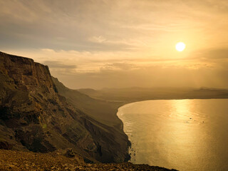 Dramatic coastal landscape unfolds with towering dark cliffs descending into the misty ocean, where a sliver of sun breaks through the clouds. Vast expanse of water and land creates a majestic scenery